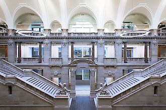 Historische Eingangshalle des Palais de Justice in Straßburg. Zwei breite Treppen führen symmetrisch nach oben zu einer Balustrade, die mit detailreichen Steinmetzarbeiten verziert ist. Auf den Podesten am Fuß der Treppen stehen kunstvolle Sphinxstatuen. Die Decke ist mit weißen, gewölbten Bögen gestaltet, die ein Gefühl von Höhe und Raum vermitteln. Moderne Elemente wie kreisförmige Leuchten kontrastieren elegant mit der klassischen Architektur. Im Hintergrund sind große Fenster zu sehen, die viel natürliches Licht in den Raum lassen.

