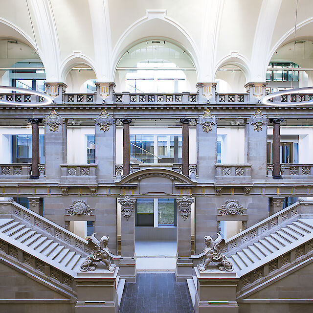 Historische Eingangshalle des Palais de Justice in Straßburg. Zwei breite Treppen führen symmetrisch nach oben zu einer Balustrade, die mit detailreichen Steinmetzarbeiten verziert ist. Auf den Podesten am Fuß der Treppen stehen kunstvolle Sphinxstatuen. Die Decke ist mit weißen, gewölbten Bögen gestaltet, die ein Gefühl von Höhe und Raum vermitteln. Moderne Elemente wie kreisförmige Leuchten kontrastieren elegant mit der klassischen Architektur. Im Hintergrund sind große Fenster zu sehen, die viel natürliches Licht in den Raum lassen.
