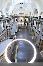 Historische Eingangshalle des Palais de Justice in Straßburg. Zwei breite Treppen führen symmetrisch nach oben zu einer Balustrade, die mit detailreichen Steinmetzarbeiten verziert ist. Auf den Podesten am Fuß der Treppen stehen kunstvolle Sphinxstatuen. Die Decke ist mit weißen, gewölbten Bögen gestaltet, die ein Gefühl von Höhe und Raum vermitteln. Moderne Elemente wie kreisförmige Leuchten kontrastieren elegant mit der klassischen Architektur. Im Hintergrund sind große Fenster zu sehen, die viel natürliches Licht in den Raum lassen.