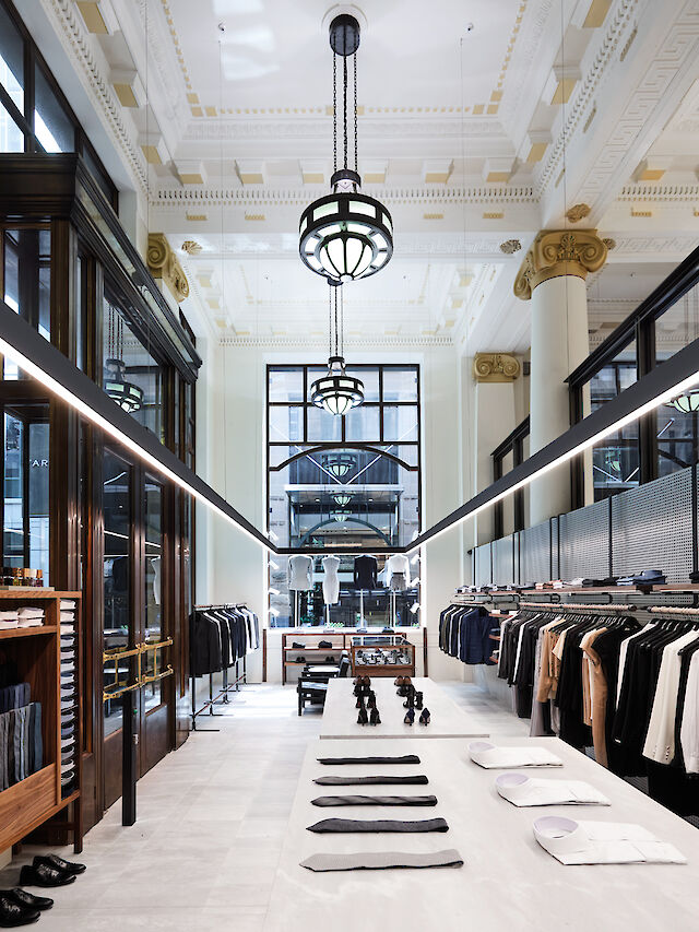 The picture shows the interior of an elegant clothing shop with high ceilings and large windows. The shop has a modern yet classic atmosphere with white walls and golden columns. In the centre of the room are tables with neatly folded shirts and ties. Above the tables are lines of light that illuminate the room well. On both sides of the room are clothes rails with various items of clothing such as jackets and shirts. Large, round ceiling lights hang from the ceiling. In the background is a large window that lets in plenty of natural light and makes the room bright and inviting.