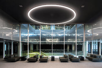 Un moderno salón con varios sofás grises y mesitas. El techo está decorado con un gran anillo iluminado. Una pared de cristal separa el salón de un atrio abierto que ocupa dos plantas. El suelo está cubierto con una alfombra de color claro y el atrio está rodeado por una galería en la planta superior.