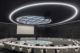 Una moderna sala de conferencias con una gran mesa redonda en el centro, rodeada de sillas blancas tapizadas. Un gran panel luminoso redondo cuelga del techo, rodeado por un anillo incandescente. Una gran pantalla cuelga de la pared del fondo.