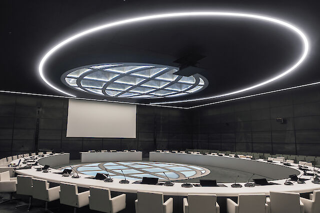 A modern conference room with a large round table in the centre, surrounded by white, upholstered chairs. A large, round light panel hangs from the ceiling, surrounded by a glowing ring. A large screen hangs on the wall in the background.
