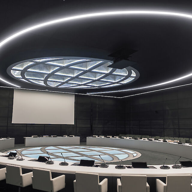Una moderna sala de conferencias con una gran mesa redonda en el centro, rodeada de sillas blancas tapizadas. Un gran panel luminoso redondo cuelga del techo, rodeado por un anillo incandescente. Una gran pantalla cuelga de la pared del fondo.