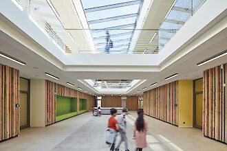 La imagen muestra el interior moderno y luminoso de un edificio con un gran techo de cristal. En primer plano, tres niños corren por el centro de la sala. Las paredes están revestidas con paneles verticales de madera y a la izquierda se ve una superficie de pared verde. Al fondo se ve una mesa con sillas. Se ve a otra persona en la galería. La sala parece abierta y acogedora, con mucha luz natural. El techo está equipado con varias luminarias lineales de superficie.