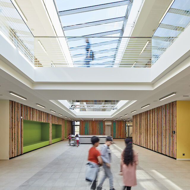 La imagen muestra el interior moderno y luminoso de un edificio con un gran techo de cristal. En primer plano, tres niños corren por el centro de la sala. Las paredes están revestidas con paneles verticales de madera y a la izquierda se ve una superficie de pared verde. Al fondo se ve una mesa con sillas. Se ve a otra persona en la galería. La sala parece abierta y acogedora, con mucha luz natural. El techo está equipado con varias luminarias lineales de superficie.