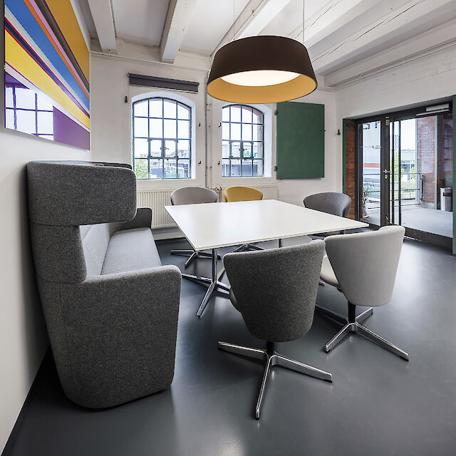 Una moderna sala de conferencias con una gran mesa rectangular blanca y cinco sillas giratorias grises alrededor. A la izquierda hay un sofá gris de respaldo alto. En la pared, detrás del sofá, cuelga una colorida obra de arte con rayas horizontales de distintos colores. Dos grandes ventanales con marcos negros dejan entrar mucha luz natural. Sobre la mesa cuelga una gran lámpara colgante redonda con una amplia pantalla negra y un cálido interior amarillo. A la derecha, una puerta de cristal da a una terraza. Las paredes son blancas y el suelo gris oscuro.