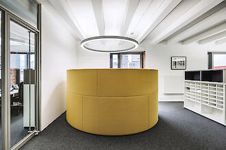 Un espacio de oficina moderno con un llamativo asiento redondo de color amarillo en el centro de la sala, con forma de pared a media altura. Sobre el asiento cuelga una gran lámpara colgante circular de color plateado. Las paredes son blancas y el techo blanco con vigas a la vista. En el lado derecho de la sala hay estanterías blancas con compartimentos abiertos. Dos ventanas dejan pasar la luz del día y a la izquierda hay una puerta de cristal que conduce a otra zona de oficinas. El suelo está cubierto de moqueta gris oscura.