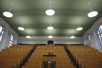 The picture shows an empty lecture theatre at Humboldt University in Berlin with wooden seats arranged in ascending rows. Several round, white pendant luminaires from the Basic Max series by Lightnet hang from the green ceiling. The walls have large windows that let in natural light. The room has a light wall colour. 