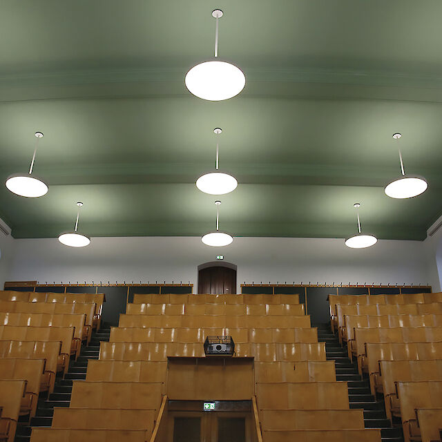 Das Bild zeigt einen leeren Hörsaal in der Humboldt-Universität Berlin mit Holzsitzen, die in ansteigenden Reihen angeordnet sind. An der grünen Decke hängen mehrere runde, weiße Pendelleuchten der Serie Basic Max von Lightnet. Die Wände haben große Fenster, durch die natürliches Licht einfällt. Der Raum hat eine helle Wandfarbe. 
