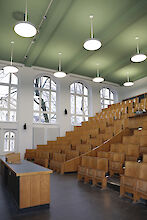 The picture shows an empty lecture theatre at Humboldt University in Berlin with wooden seats arranged in ascending rows. Several round, white pendant luminaires from the Basic Max series by Lightnet hang from the green ceiling. The walls have large windows that let in natural light. The room has a light wall colour. A desk can be seen at the bottom of the picture.