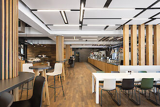 Restaurante con elementos de madera, mobiliario en blanco y negro y luminarias lineales en el techo.