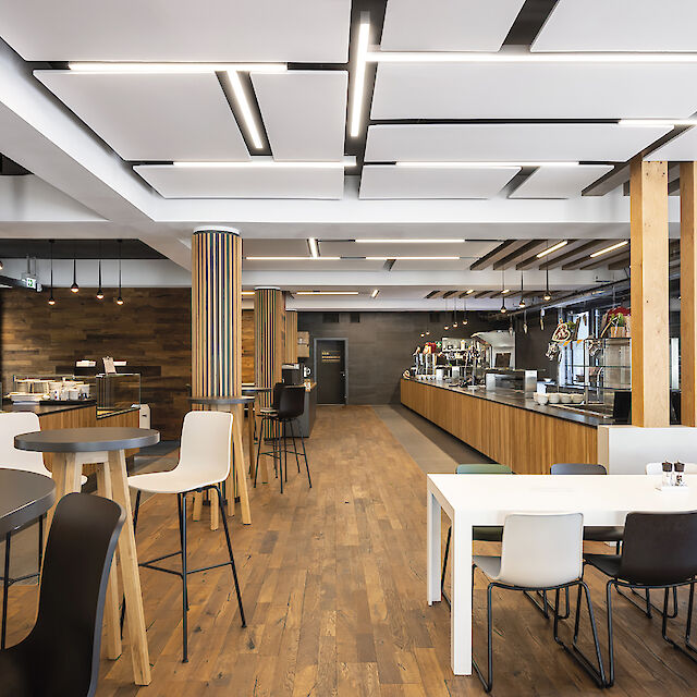 Restaurante con elementos de madera, mobiliario en blanco y negro y luminarias lineales en el techo.
