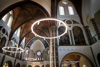 Historische Kirchendecke mit Bögen, bunten Glasfenstern und modernen, ringförmigen LED-Pendelleuchten.