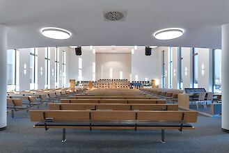 Iglesia moderna con ventanas altas, bancos de madera y luminarias redondas LED de superficie.