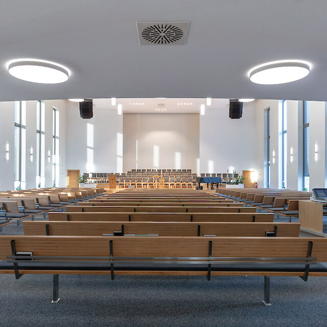 Iglesia moderna con ventanas altas, bancos de madera y luminarias redondas LED de superficie.