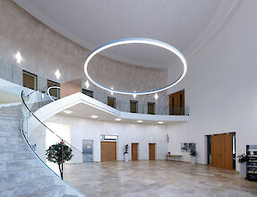 Amplio vestíbulo con escalera curva, detalles de madera y una gran luminaria LED suspendida en forma de anillo.