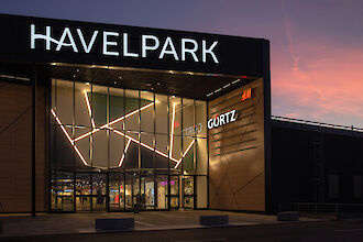 Der Haupteingang des Einkaufszentrums Havelpark Dallgow bei Sonnenuntergang, beleuchtet mit modernen Lichtstrukturen. Die Fassade besteht aus großen Glasflächen, die von einem geometrischen Muster aus Lichtakzenten durchzogen sind, und wird von hölzernen Elementen flankiert. Das beleuchtete Logo 
