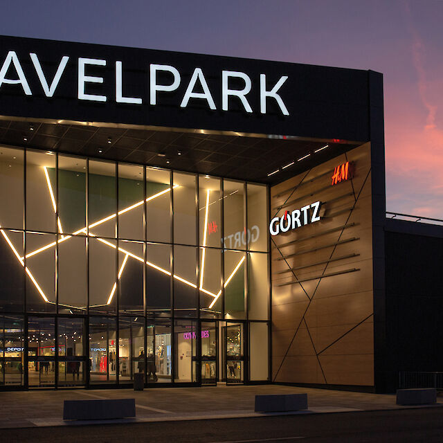 La entrada principal del centro comercial Havelpark Dallgow al atardecer, iluminada con modernas estructuras de luz. La fachada está formada por grandes superficies de cristal intercaladas con un patrón geométrico de acentos luminosos y flanqueada por elementos de madera. El logotipo 