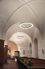 Iglesia medieval con paredes de ladrillo blanco, bancos de madera y luminarias LED suspendidas en forma de anillo.