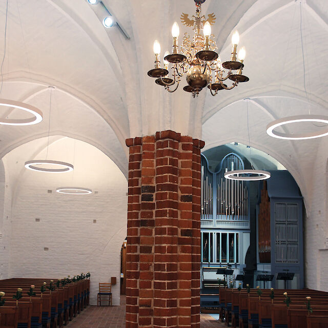 Iglesia histórica con techo abovedado blanco, columna de ladrillo, lámpara de araña y modernas luminarias suspendidas LED en forma de anillo.