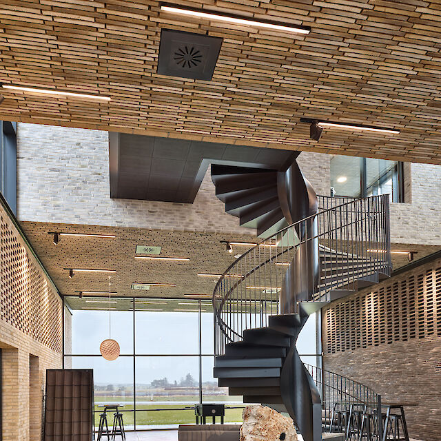 La imagen muestra el interior de un edificio moderno con un diseño abierto y aireado. La pieza central es una elegante escalera de caracol de metal negro que asciende desde la planta baja. Las paredes y el techo están revestidos de ladrillos rectangulares claros, que dan a la estancia un ambiente cálido y acogedor. En el techo hay varias lámparas lineales de superficie que iluminan uniformemente la sala. Al fondo, grandes ventanales dejan entrar mucha luz natural y ofrecen vistas a un paisaje verde. En la parte delantera de la sala hay una gran piedra que sirve de decoración, así como algunos asientos y una mesa de bar con taburetes.