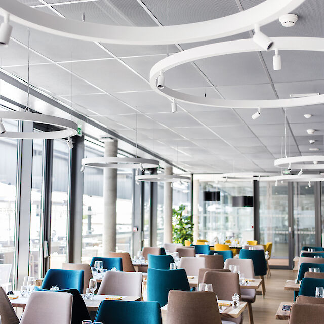 Restaurante con grandes ventanales, mobiliario elegante y luminarias suspendidas LED en forma de anillo con focos.