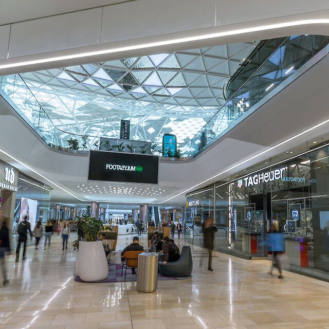 Innenansicht eines modernen Einkaufszentrums mit gläsernem Dach. Es gibt mehrere Geschäfte auf beiden Seiten. Im Zentrum befinden sich Sitzgelegenheiten und Pflanzen in großen weißen Pflanzgefäßen. Einige Personen gehen durch die Gänge, während andere vor den Geschäften stehen. Die Beleuchtung ist hell und das Ambiente wirkt futuristisch und einladend. An der Decke befindet sich eine rund umlaufende Lichtlinie.