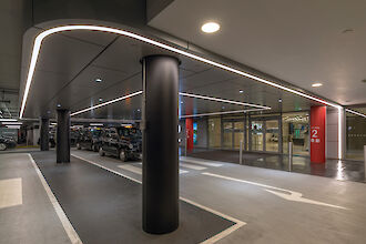 Parkhaus mit moderner, hell beleuchteter Decke und glatten, dunklen Säulen. An der Decke verläuft eine durchgehende Lichtlinie, die die Fläche gleichmäßig ausleuchtet. Mehrere schwarze Autos sind parallel entlang der Fahrbahn geparkt. Der Boden ist sauber und markiert, mit deutlichen weißen Fahrbahnmarkierungen und Pfeilen. Im Hintergrund befindet sich der Eingang zu einem Gebäude, gekennzeichnet durch rote Säulen mit der Aufschrift 'Eingang 2'.