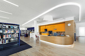 La imagen muestra el interior de una biblioteca moderna. En primer plano hay un mostrador de información y préstamo de madera curvada con dos ordenadores y una pared trasera naranja. A la izquierda hay estanterías altas con libros. El suelo es de madera clara y sobre el mostrador hay una lámpara blanca de superficie y forma cónica. Al fondo se extienden otras estanterías con libros.