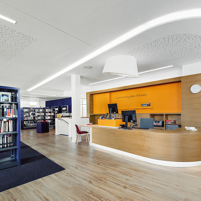 La imagen muestra el interior de una biblioteca moderna. En primer plano hay un mostrador de información y préstamo de madera curvada con dos ordenadores y una pared trasera naranja. A la izquierda hay estanterías altas con libros. El suelo es de madera clara y sobre el mostrador hay una lámpara blanca de superficie y forma cónica. Al fondo se extienden otras estanterías con libros.