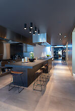 Cocina moderna iluminada por focos de superficie con mobiliario oscuro, gran isla de cocción y taburetes en la barra.