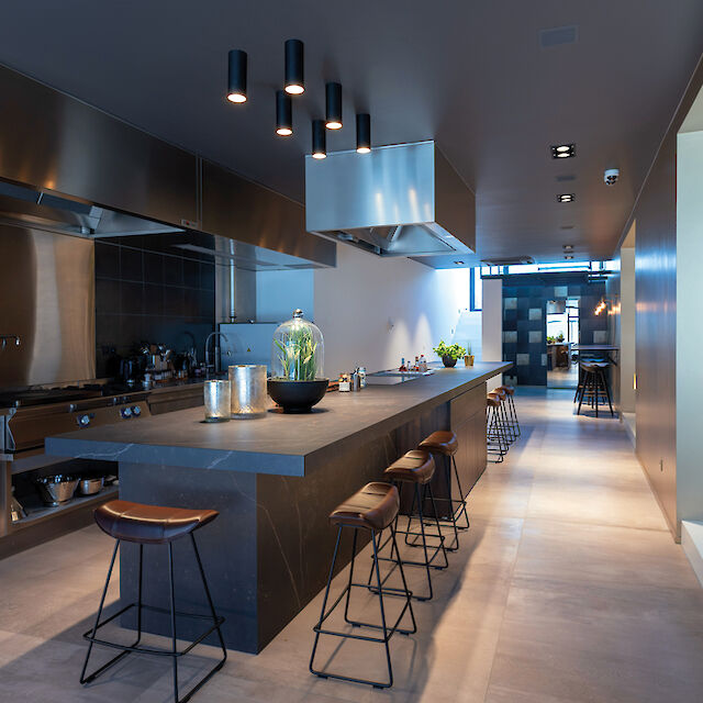 Cocina moderna iluminada por focos de superficie con mobiliario oscuro, gran isla de cocción y taburetes en la barra.