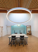 Large, ring-shaped LED pendant luminaire above a rectangular meeting table in a room with a wooden ceiling.