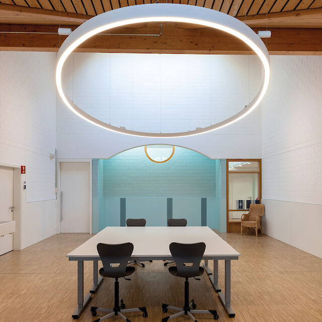Luminaria LED suspendida de gran tamaño y forma anular sobre una mesa de reuniones rectangular en una sala con techo de madera.