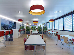 Cantina con mesas blancas, sillas de colores y luminarias suspendidas rojas Beam Me Up