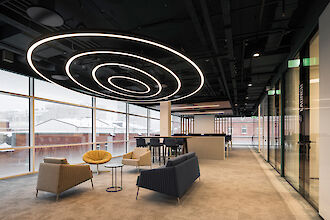 Das Bild zeigt ein modernes Büro mit großen Fenstern, die einen Blick auf verschneite Gebäude draußen bieten. An der Decke hängt eine auffällige, spiralförmige Lichtstruktur aus mehreren ovalen Pendelleuchten unterschiedlicher Größe. Im Vordergrund befinden sich mehrere Sitzgelegenheiten: ein gelber Sessel, ein beiger Sessel, eine dunkelblaue Couch und ein kleiner runder Beistelltisch.