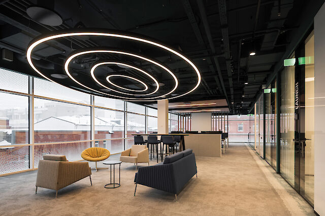 The image shows a modern office with large windows looking out onto snow-covered buildings outside. From the ceiling hangs a striking spiral light structure. It consists of several oval pendant lights of different sizes. In the foreground are several pieces of seating furniture: a yellow armchair, a beige armchair, a dark blue couch and a small round side table.