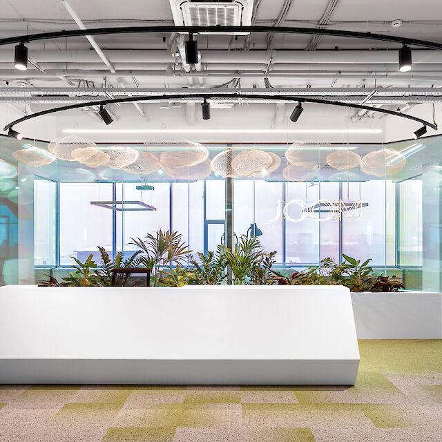 La imagen muestra la recepción de una oficina moderna con un gran mostrador blanco en primer plano. Detrás del mostrador hay una zona con numerosas plantas y perfiles negros de aluminio curvado con focos LED negros, que crean un ambiente cálido y acogedor. La pared trasera es de cristal, a través de la cual se ven las oficinas. El techo es abierto, con tuberías y cables a la vista, lo que da al espacio un aire industrial. El suelo está cubierto de losetas de moqueta verde, que crean una impresión fresca y viva.