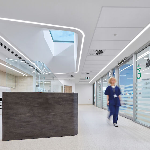 Pasillo de hospital con líneas de luz LED de forma orgánica en el techo, mostrador de recepción y cuidador ambulante