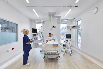 Luminosa habitación de hospital con paciente en la cama, equipo médico y luminarias empotradas lineales en el techo.