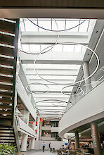 Ein modernes Atrium in einem Gebäude mit einem hohen Glasdach, das viel natürliches Licht hereinlässt. Von der Decke hängen mehrere große, ringförmige Leuchten in verschiedenen Größen, die in einem kunstvollen Muster angeordnet sind. Auf der linken Seite des Bildes führt eine breite Treppe nach oben. Unten rechts im Bild ist ein Café mit Sitzbereichen. Menschen bewegen sich durch den offenen Raum, der durch seine helle und moderne Gestaltung auffällt.