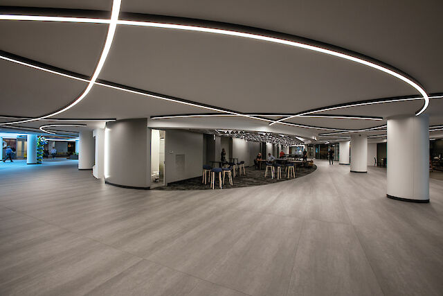 A modern, spacious interior of a building with an open-plan layout. The ceiling is designed with elegant, curved LED light lines that form a futuristic pattern. The floor is made of light grey laminate and there are several tables and chairs in the middle of the room. The walls are minimalist and there are large, round columns dividing the room. Seating areas can be seen in the background.