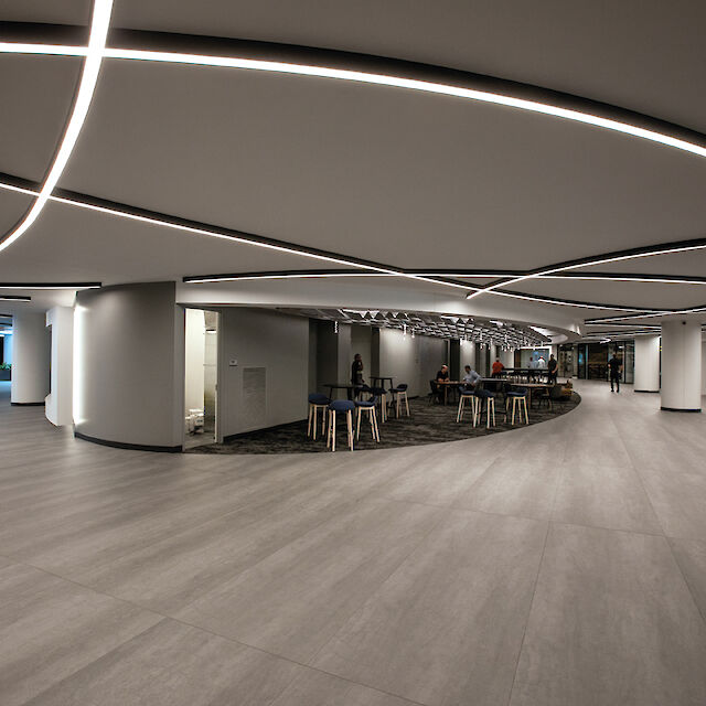 Ein moderner, großzügiger Innenraum eines Gebäudes mit einem offenen Grundriss. Die Decke ist mit eleganten, geschwungenen LED-Lichtlinien gestaltet, die ein futuristisches Muster bilden. Der Boden besteht aus hellgrauem Laminat, und in der Mitte des Raumes befinden sich mehrere Tische und Stühle. Die Wände sind minimalistisch gehalten, und es gibt große, runde Säulen, die den Raum unterteilen. Im Hintergrund sind Sitzbereiche zu sehen.