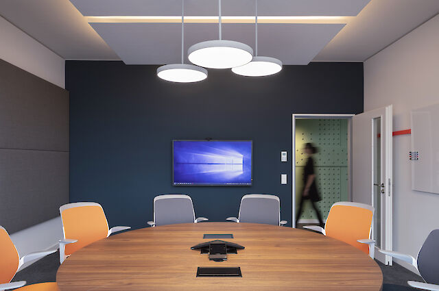 A modern conference room with a round wooden table and several chairs in grey and orange. Three round pendant lights hang from the ceiling, illuminating the room evenly. A large screen with the Windows start screen hangs on the dark blue wall opposite the table. A person walks through an open door in the background. There is a whiteboard on the right-hand wall.