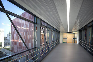 Corridor with glass façade, metal struts and linear ceiling lighting, with a view of a modern building.