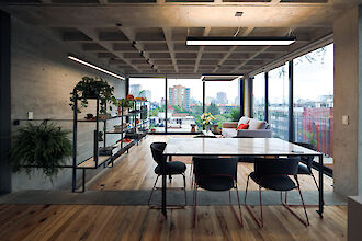 Bright living room with large window fronts, plants, dining table and rectangular pendant lights on concrete ceiling