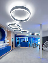 Moderna zona de recepción de diseño futurista. Del techo cuelgan luces LED luminosas en forma de anillo con patrones curvos. A la izquierda hay una pared azul con estantes en los que se exponen toallas blancas y productos de cuidado. El mostrador de recepción es azul y semicircular, con torniquetes de acceso detrás. El suelo está cubierto de baldosas de color gris claro. Una mecedora esférica blanca cuelga del techo a la derecha de la imagen. 





