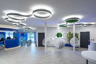 Moderna recepción diáfana de diseño futurista. El techo está decorado con luces LED luminosas en forma de anillo dispuestas en patrones orgánicos interconectados. Algunas de las luces contienen inserciones de musgo. En primer plano, tres mecedoras esféricas blancas cuelgan del techo. El suelo está cubierto de baldosas de color gris claro. A la izquierda de la imagen hay un mostrador de recepción de color azul moderno, detrás del cual hay un torniquete de acceso.