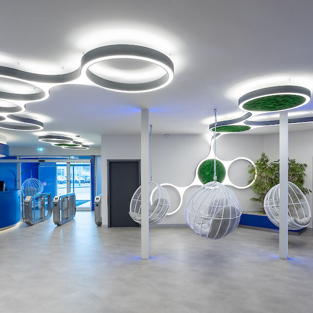 Moderner, offener Empfangsbereich mit einem futuristischen Design. Die Decke ist mit ringförmigen, leuchtenden LED-Leuchten verziert, die in organischen, miteinander verbundenen Mustern angeordnet sind. Einige der Leuchten enthalten Moos-Einsätze. Im Vordergrund hängen drei weiße, kugelförmige Schaukelstühle von der Decke. Der Boden ist mit hellgrauen Fliesen ausgelegt. Links im Bild befindet sich ein Empfangstresen in modernem Blau, hinter dem sich ein Zugangsdrehkreuz befindet.